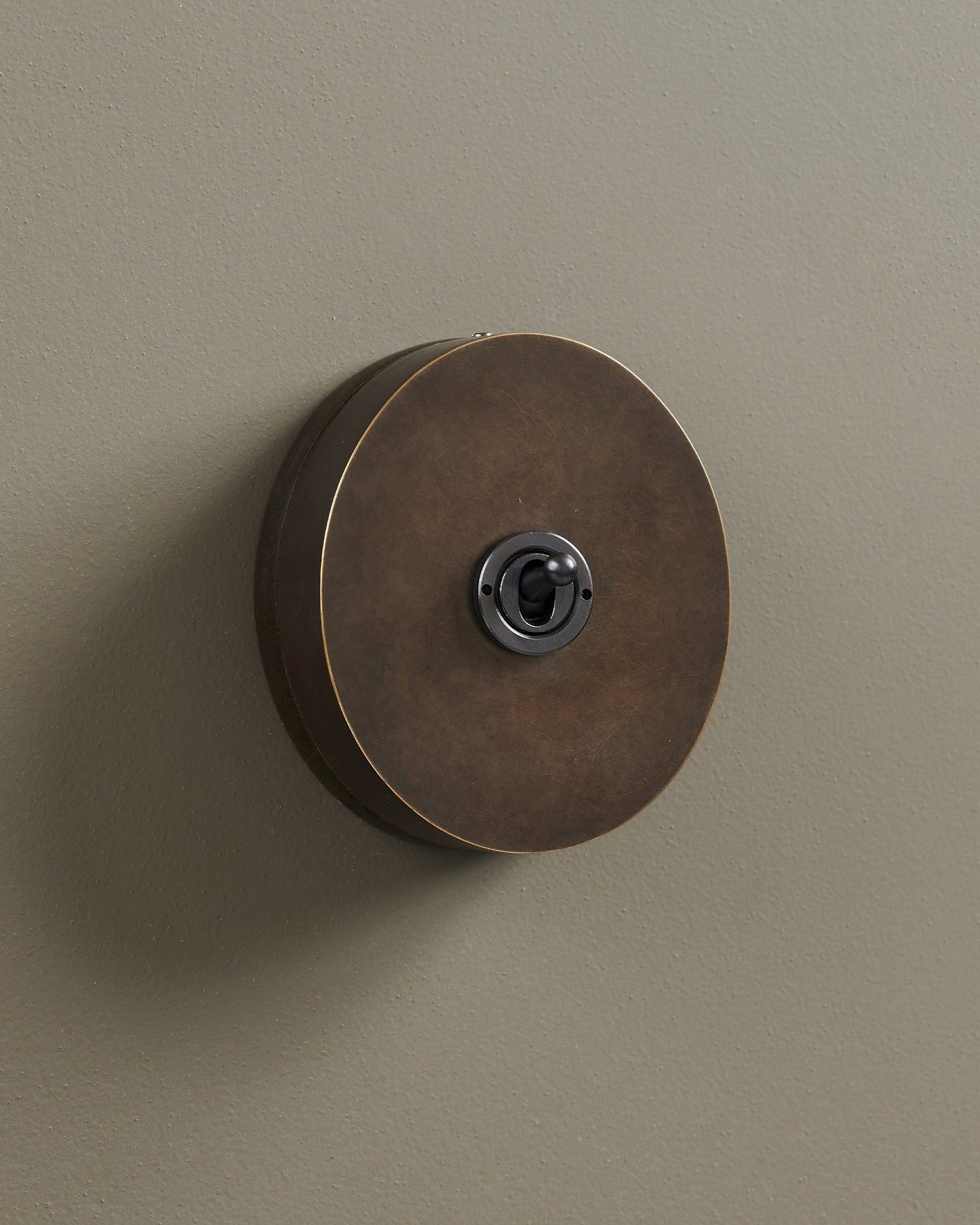 An oblique view of a 1 gang oval-shaped toggle switch in a dark oxidised brass finish with a black base.