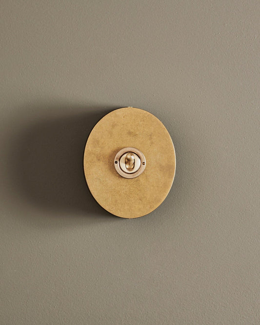 A straight-on view of a small 1 gang oval-shaped toggle switch in a mottled aged brass finish with a black base.
