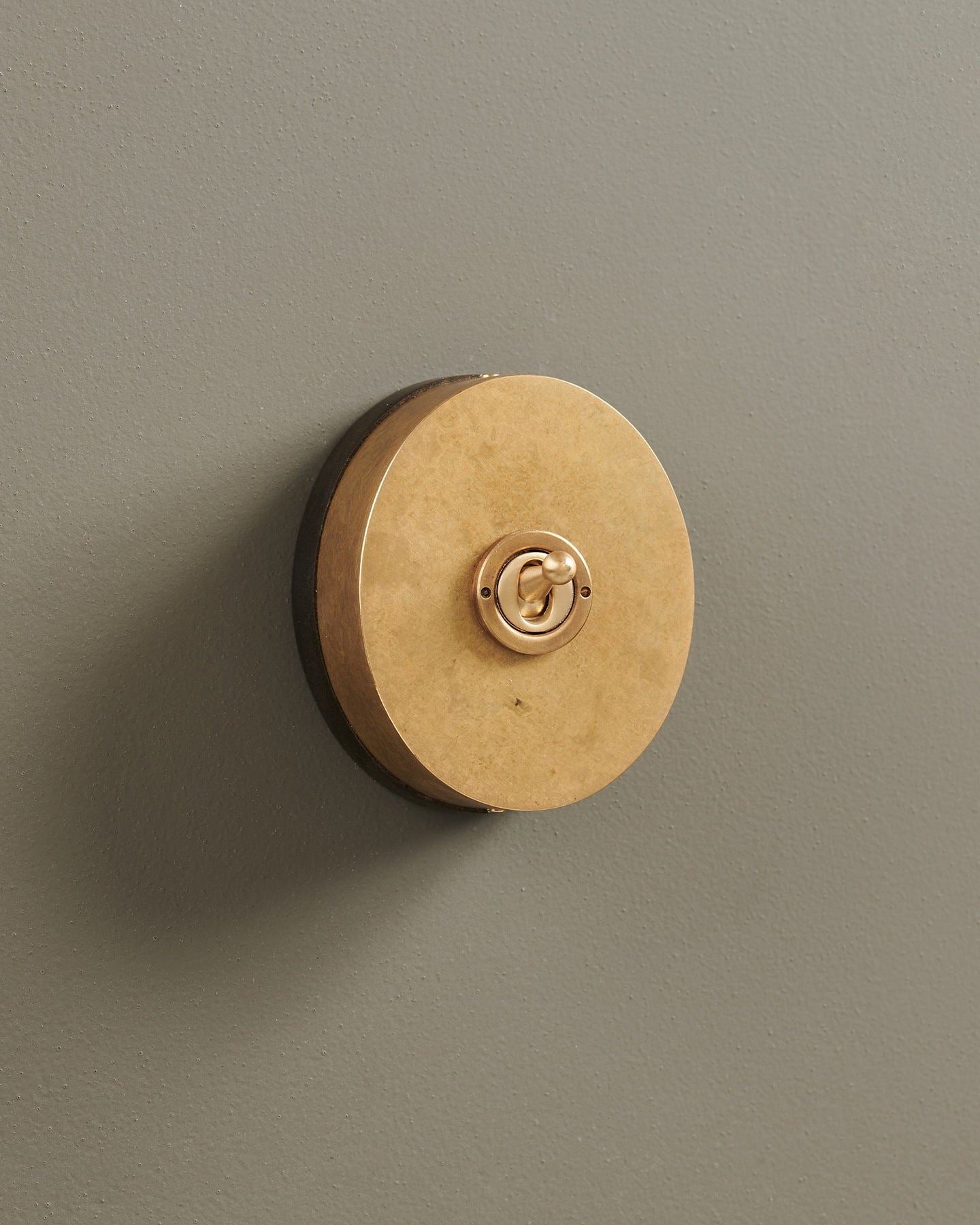 An oblique view of a small 1 gang oval-shaped toggle switch in a mottled aged brass finish with a black base.