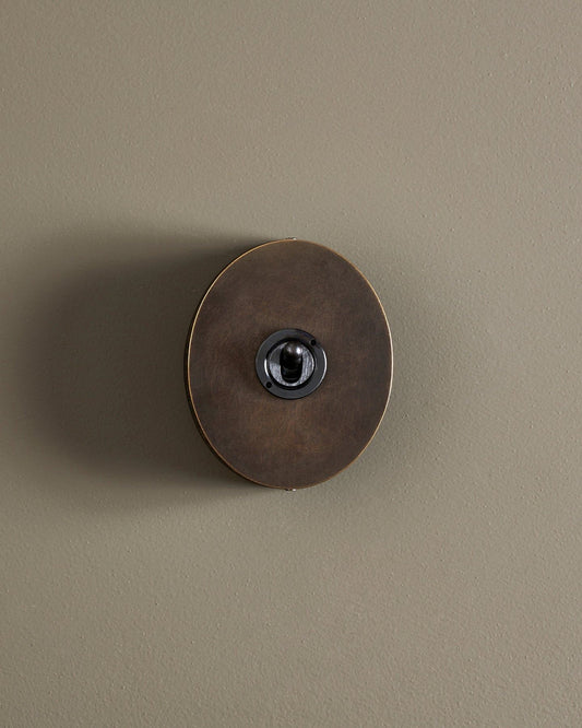 A straight-on view of a small 1 gang oval-shaped toggle switch in a dark oxidised brass finish with a black base.