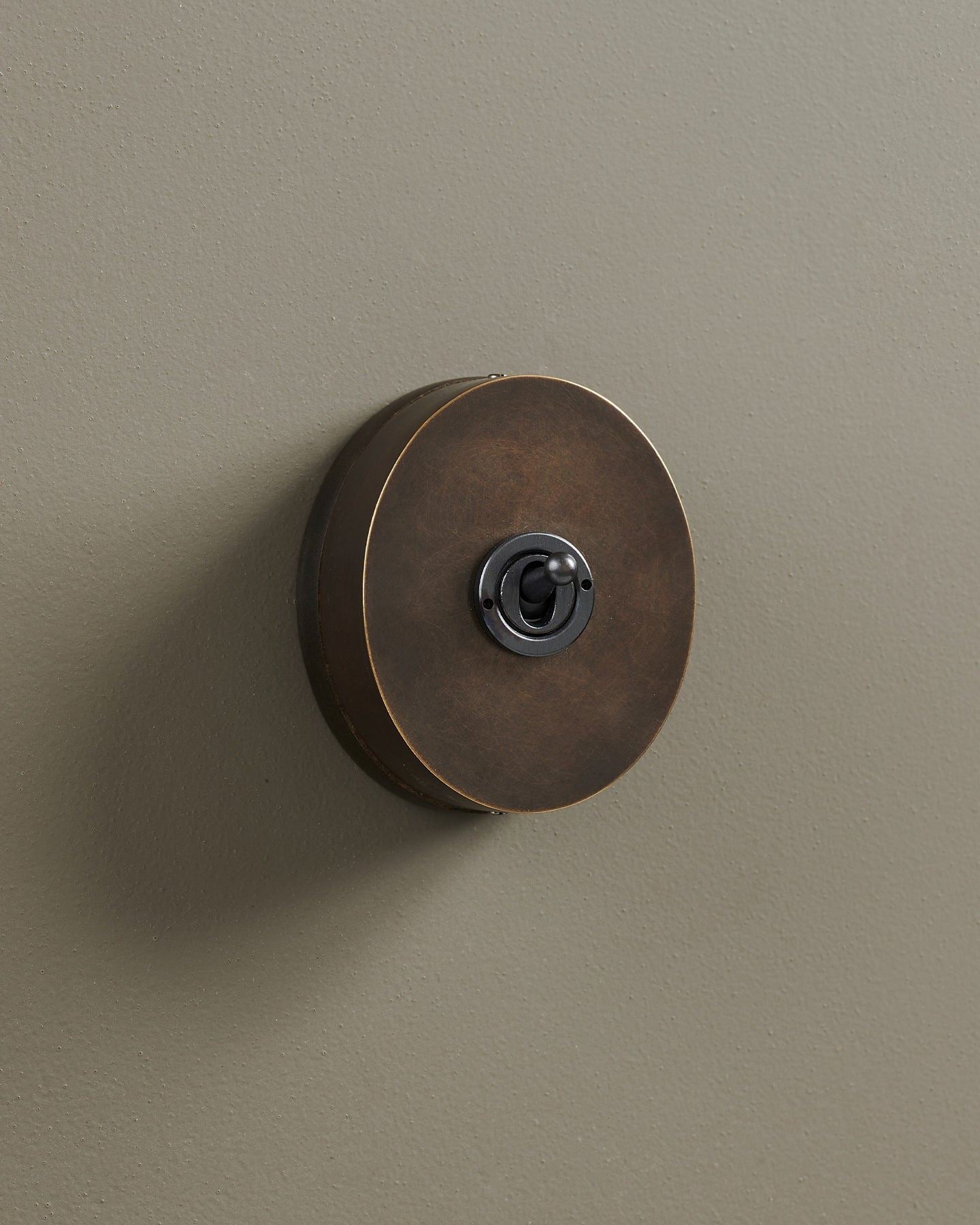 An oblique view of a small 1 gang oval-shaped toggle switch in a dark oxidised brass finish with a black base.