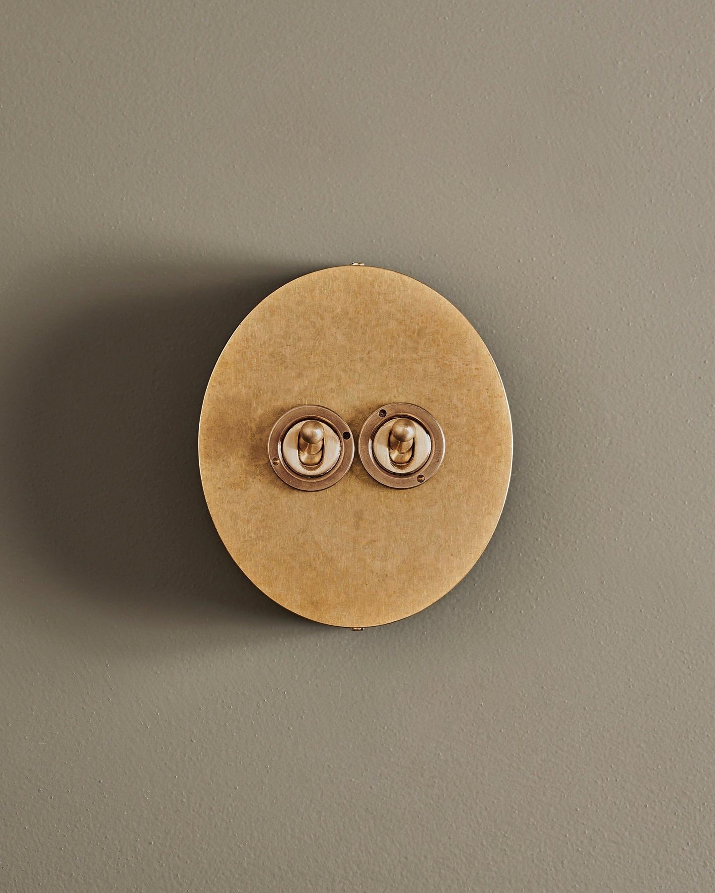 A straight-on view of a 2 gang oval-shaped toggle switch in a mottled aged brass finish with a black base.