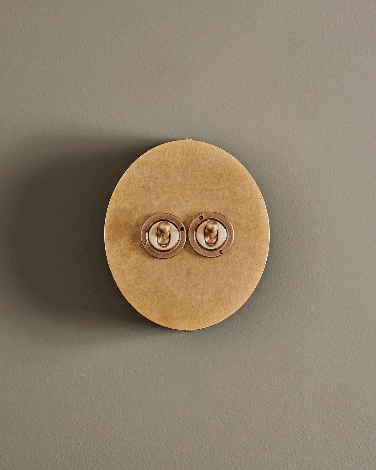 A straight-on view of a 2 gang oval-shaped toggle switch in a mottled aged brass finish with a black base. TEST