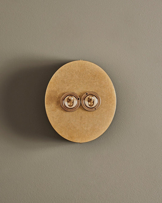A straight-on view of a 2 gang oval-shaped toggle switch in a mottled aged brass finish with a black base.