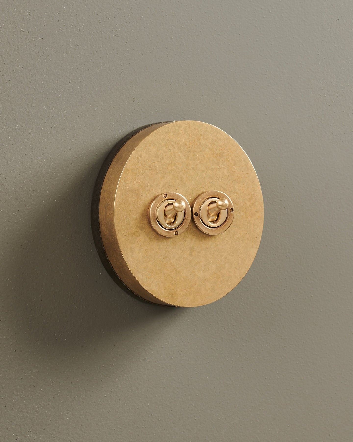 An oblique view of a 2 gang oval-shaped toggle switch in a mottled aged brass finish with a black base.