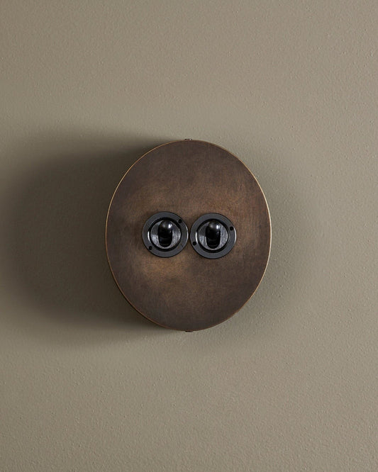 A straight-on view of a 2 gang oval-shaped toggle switch in a dark oxidised brass finish with a black base.