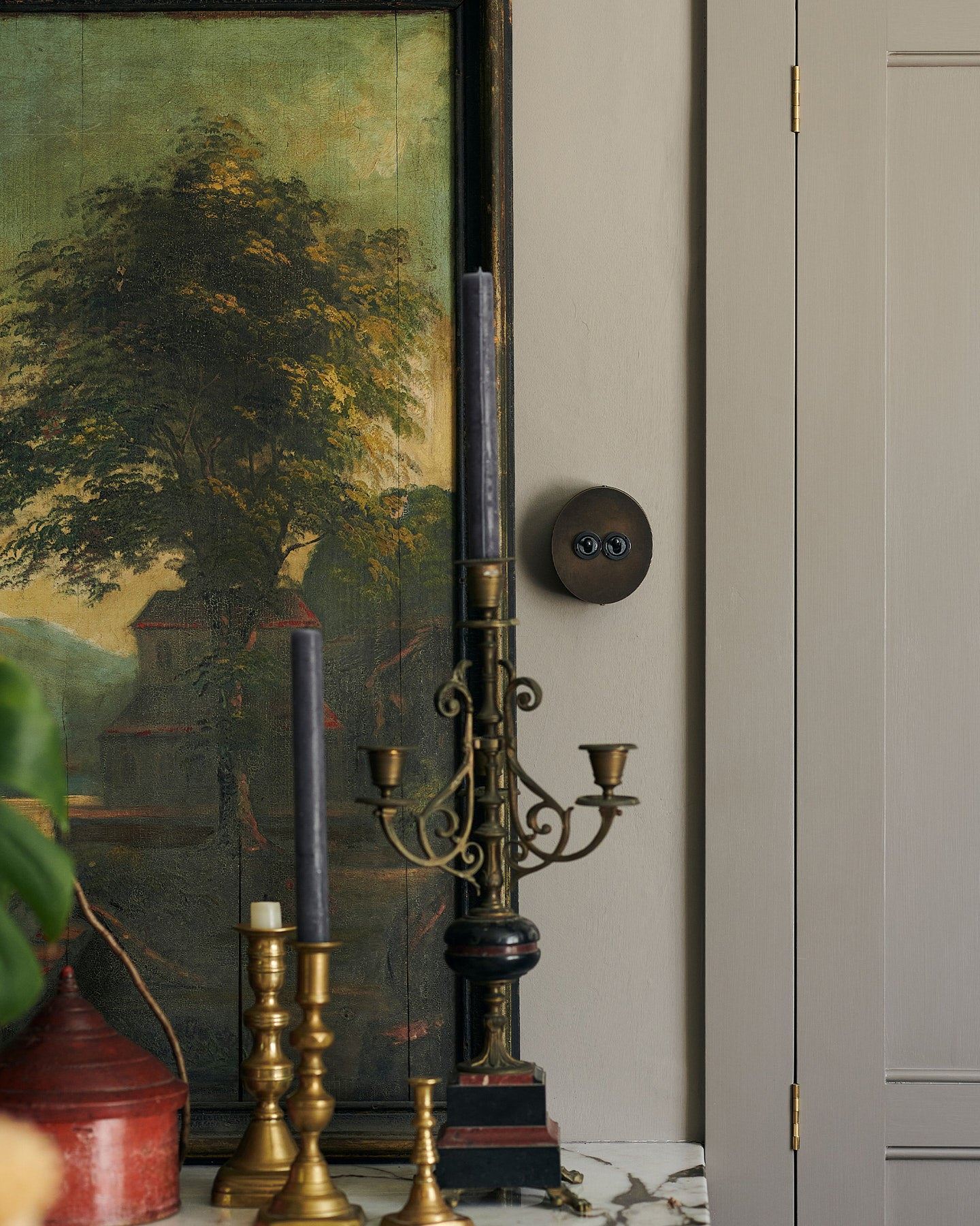 A 2 gang oval-shaped toggle switch in a dark oxidised brass finish on a grey wall, beside a large antique oil painting of a moody landscape. An elaborate tall candelabra sits in the foreground.