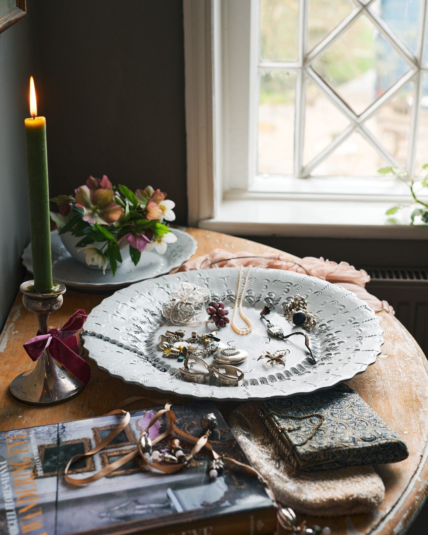 A large handmade bohemian-style glossy white ceramic platter with a scalloped rim and delicate impressions and surface patterns is covered in jewellery with a lit candle nearby.
