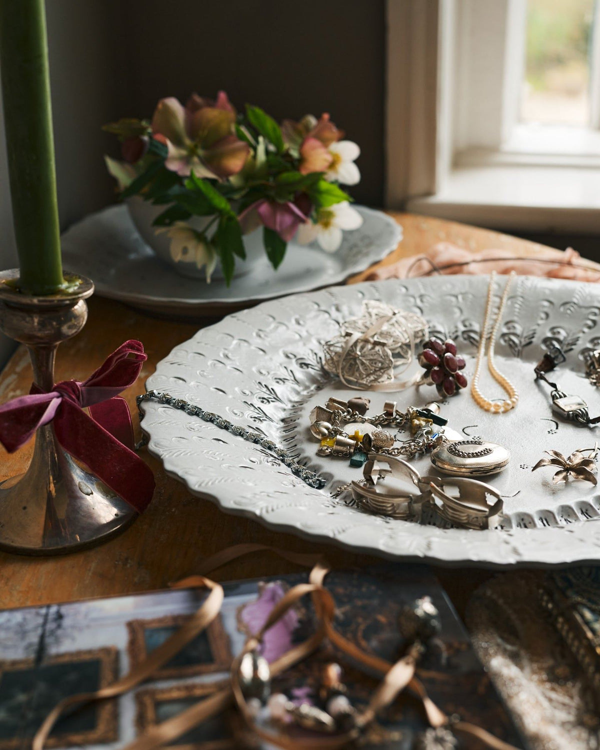 A close-up view of a large bohemian-style glossy white ceramic platter with a scalloped rim and delicate impressions and surface patterns, a selection of jewellery is on the platter and table surface. TEST