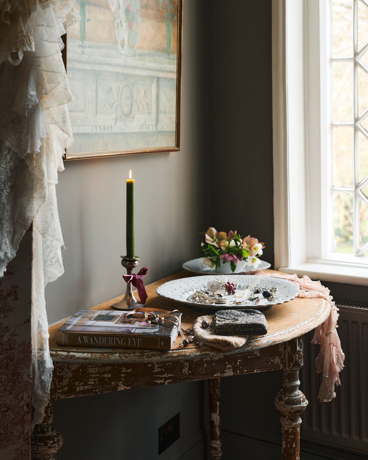 An oblique view of an antique half-moon console table by a leaded window. An antique oil painting hangs above the table, on the top is a large bohemian-style white ceramic platter, an interior design book, a lit candle and flowers.