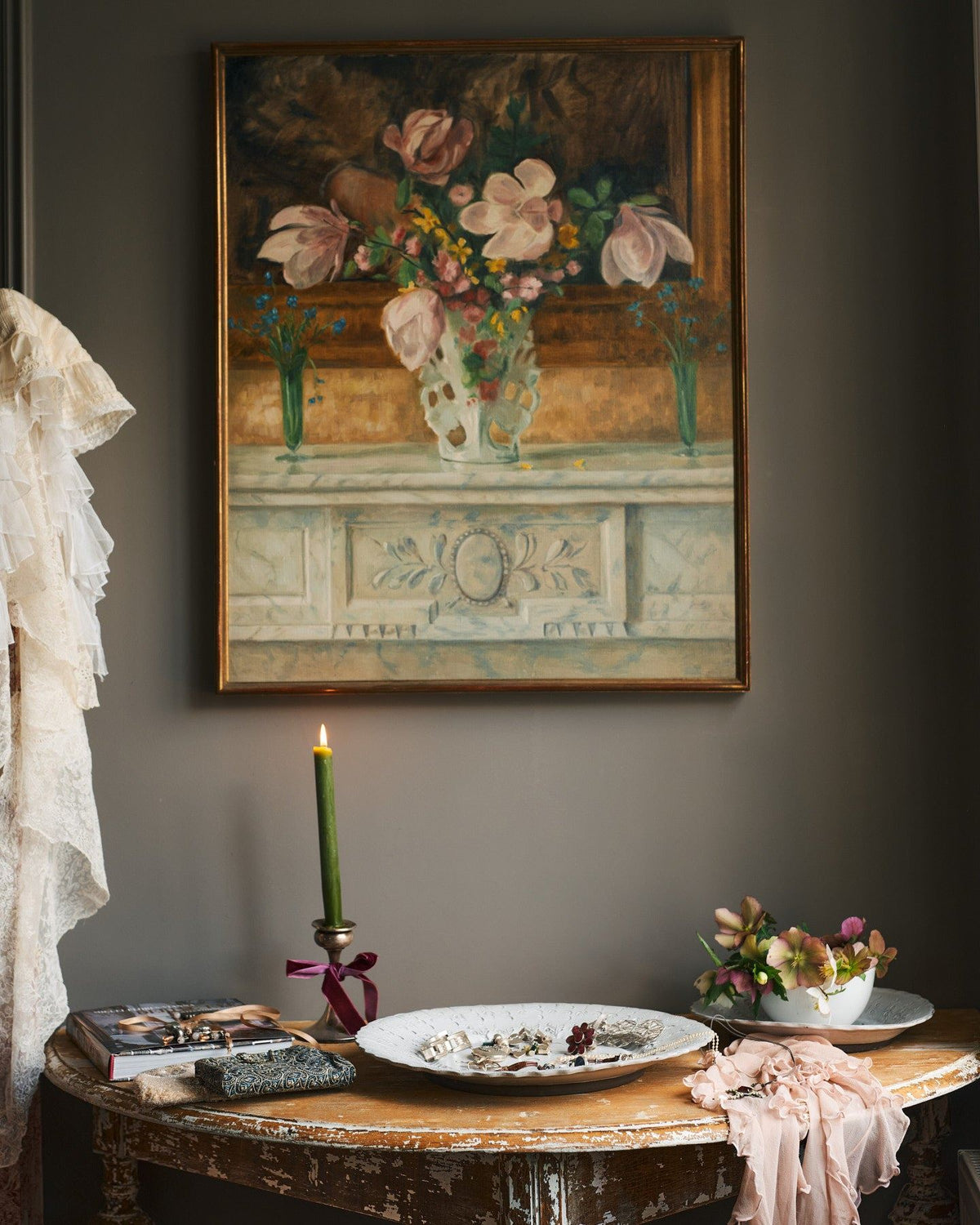 A straight-on view of an antique half-moon console table with a large antique oil painting of flowers hanging above. On the top is a large bohemian-style white ceramic platter, an interior design book, a lit candle and flowers. TEST