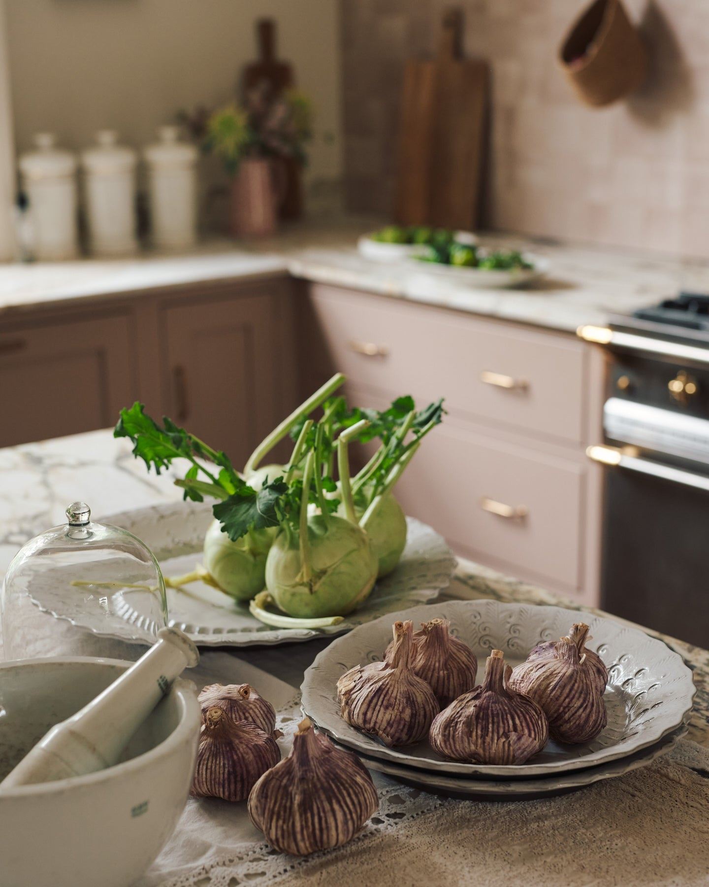 Kohlrabi and garlic heads on a large handmade bohemian-style glossy white ceramic platter with a scalloped rim, in a dusky pink Shaker-style kitchen.