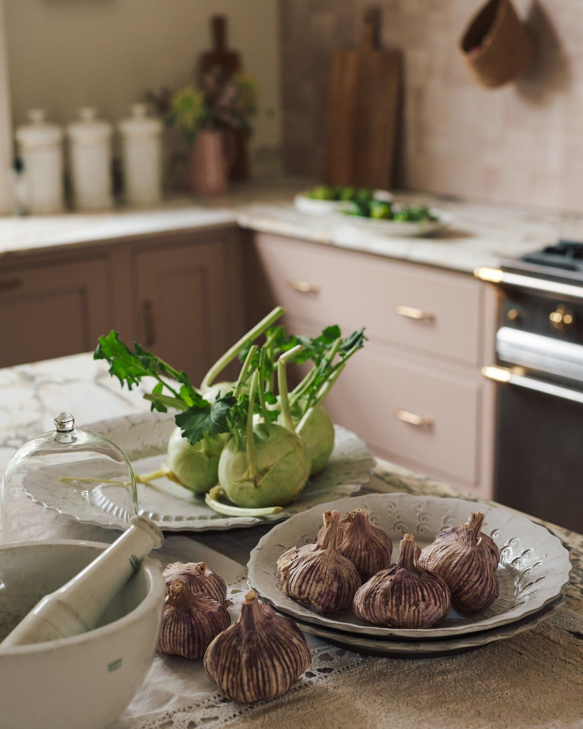 Kohlrabi and garlic heads on a large handmade bohemian-style glossy white ceramic platter with a scalloped rim, in a dusky pink Shaker-style kitchen. TEST