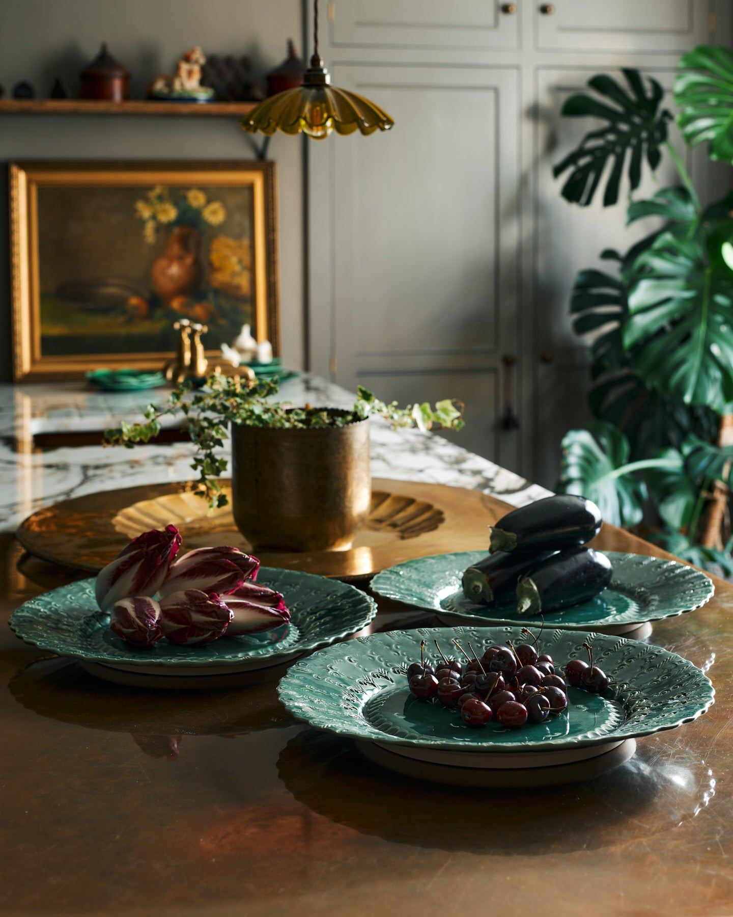 Three large handmade bohemian-style green ceramic platters with an assortement of aubergines, red chicory and cherries in an eclectic kitchen with an aged copper worktop, grey painted walls, antique paintings and a large monstera plant.