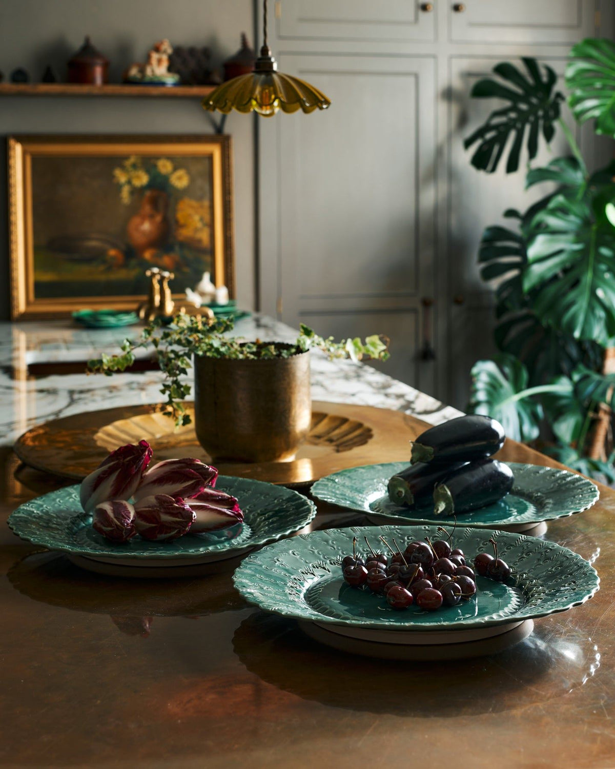 Three large handmade bohemian-style green ceramic platters with an assortement of aubergines, red chicory and cherries in an eclectic kitchen with an aged copper worktop, grey painted walls, antique paintings and a large monstera plant. TEST