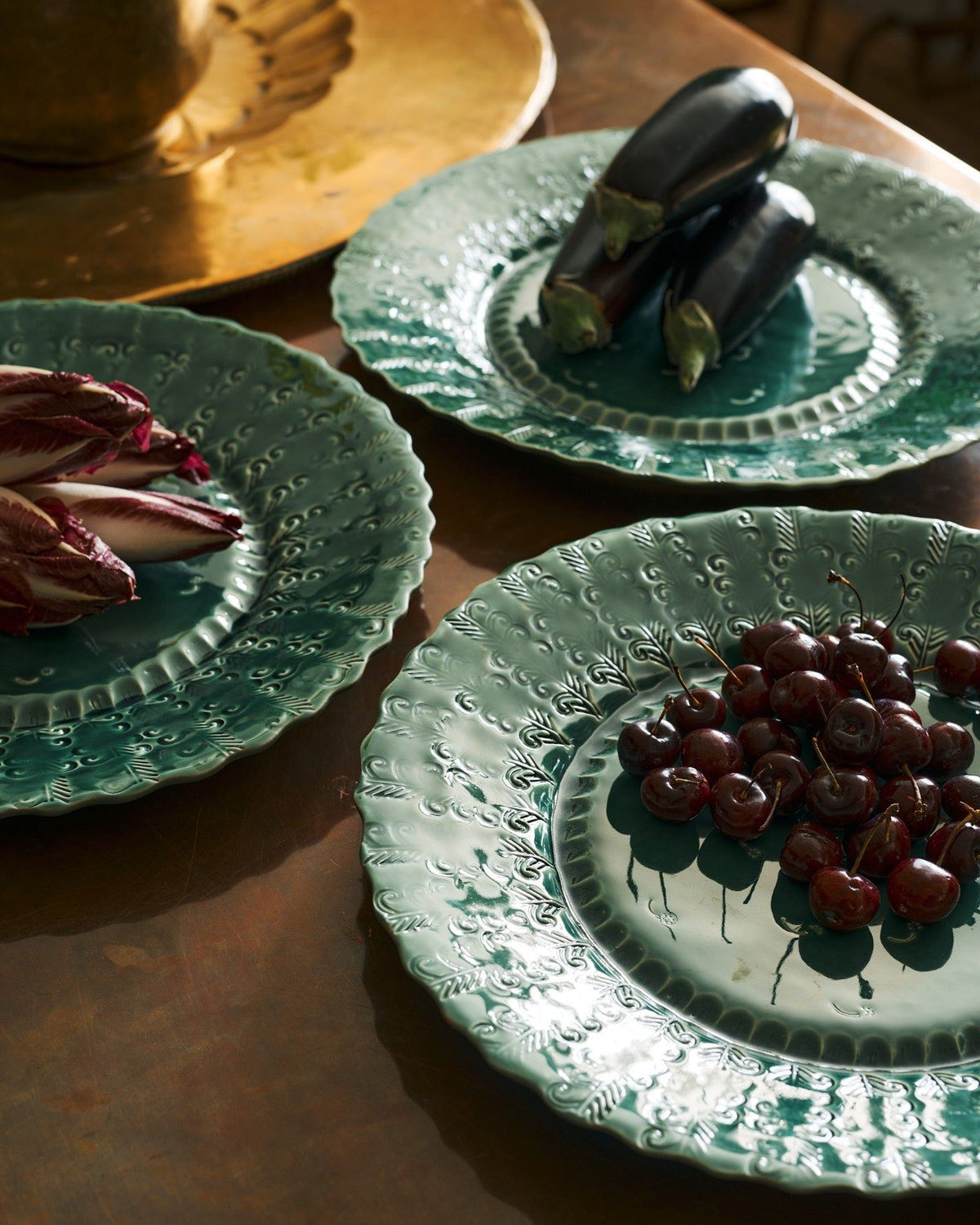 A close-up view of three large handmade bohemian-style green ceramic platters with scalloped rims and delicate impressions and surface patterns and an assortement of aubergines, red chicory and cherries. TEST