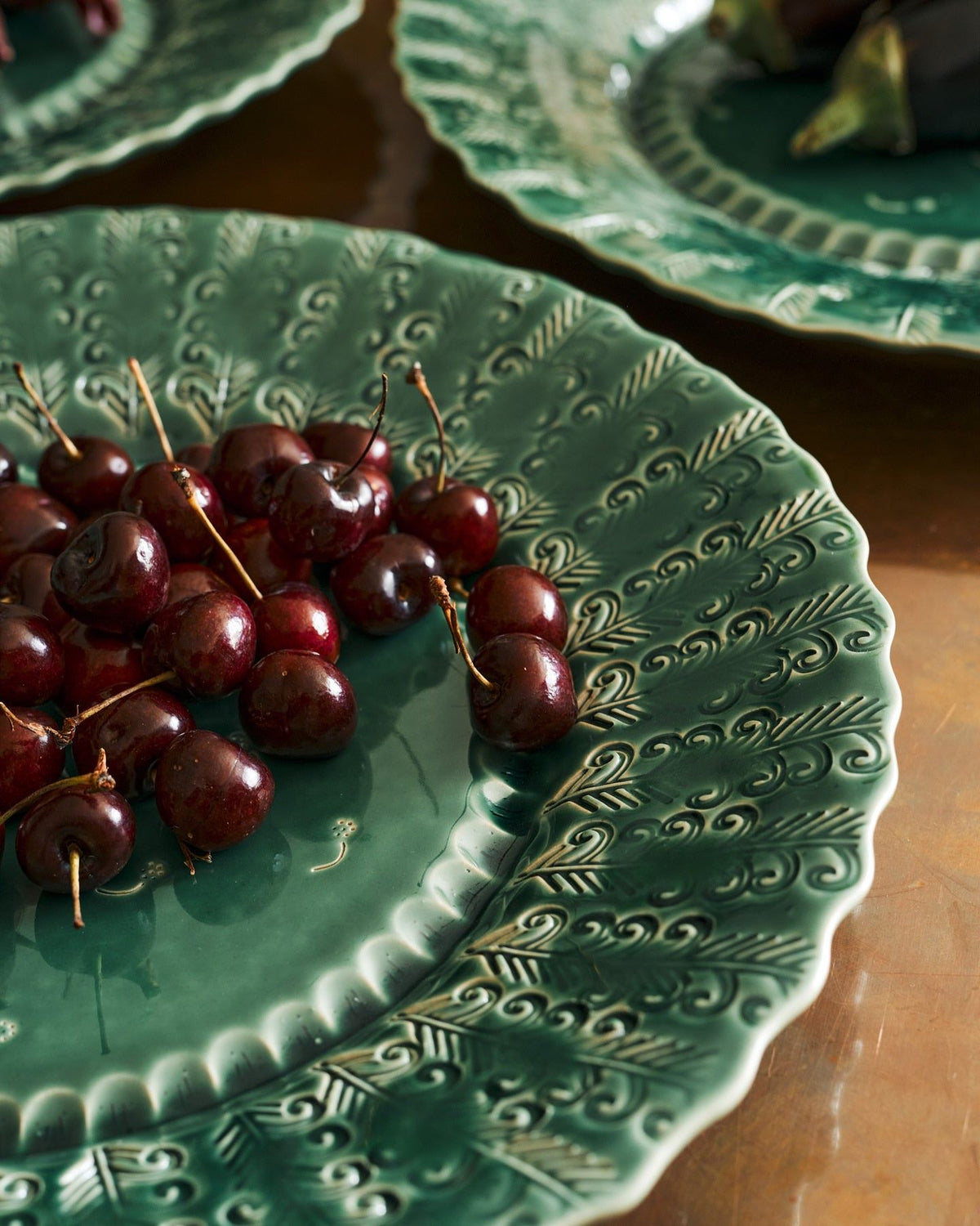 A close-up view of a bunch of cherries on a large handmade bohemian-style green ceramic platter with a scalloped rim and delicate impressions and surface patterns. TEST