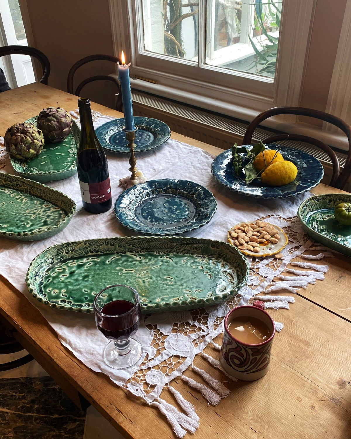 Several oval and round handmade bohemian-style ceramic platters set at a table with a vintage lace tablecloth, a bottle of red wine and a lit candle. TEST