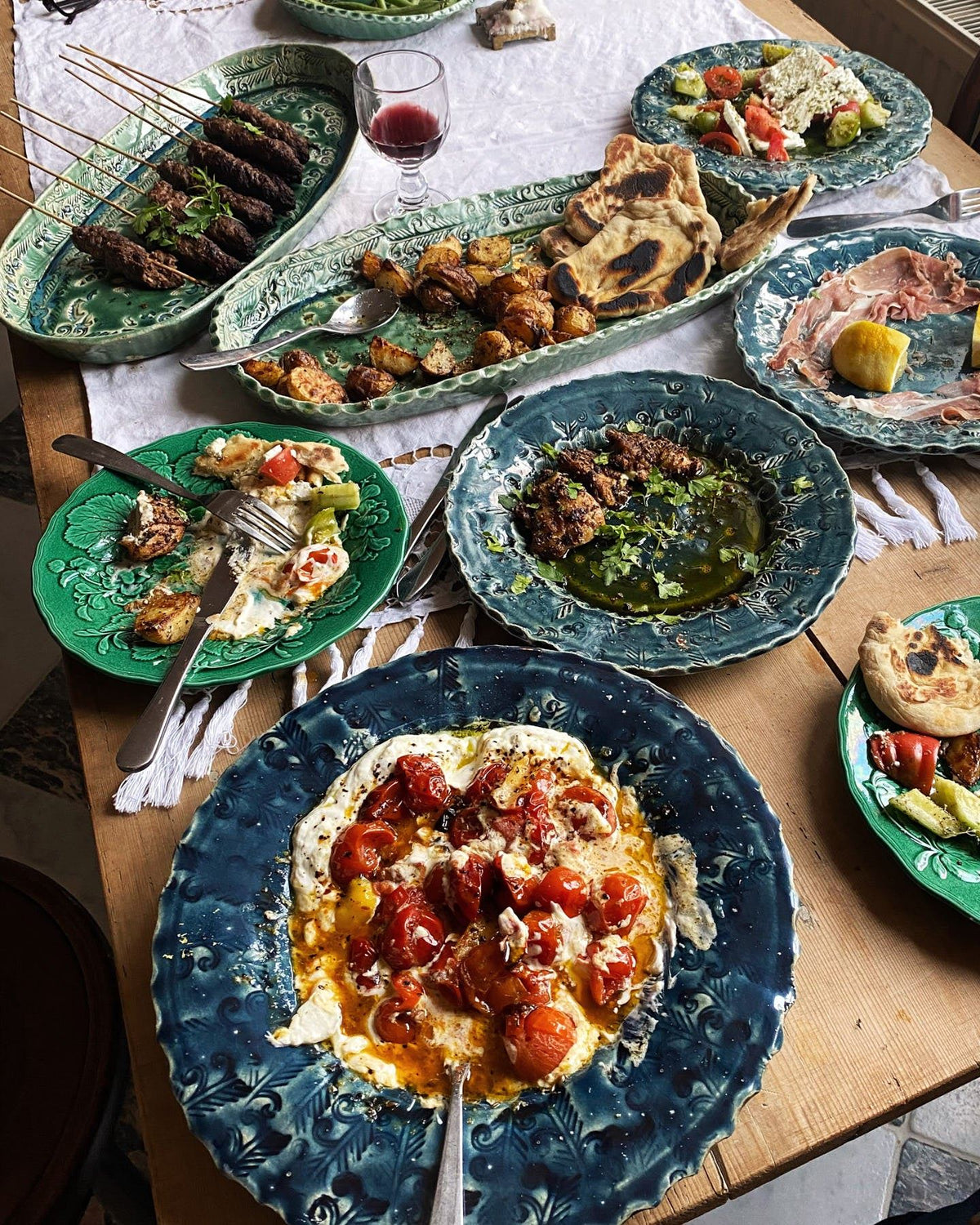 Several oval and round handmade bohemian-style ceramic platters set at a table with a vintage lace tablecloth and a variety of Mediterranean foods. TEST