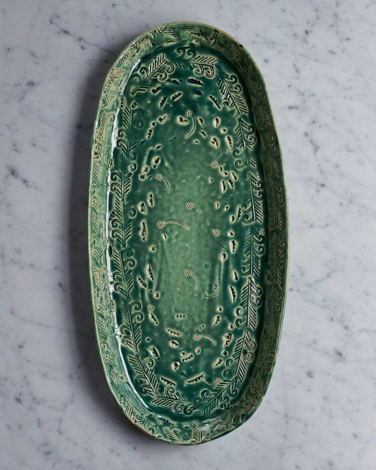 An overhead view of an oval-shaped handmade bohemian-style green ceramic platter with delicate impressions and surface patterns. TEST