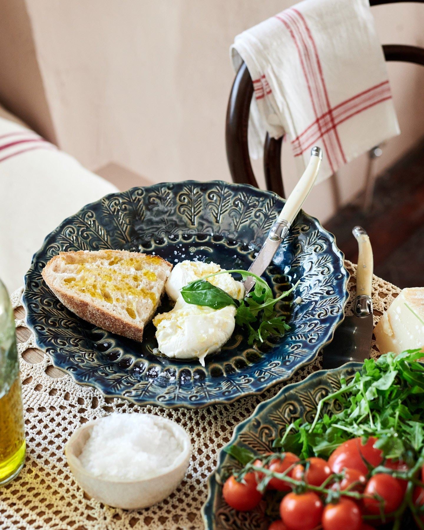A round handmade bohemian-style blue ceramic platter, with cheese, tomatoes, homemade bread and leafy greens on a beautiful crochet lace tablecloth.