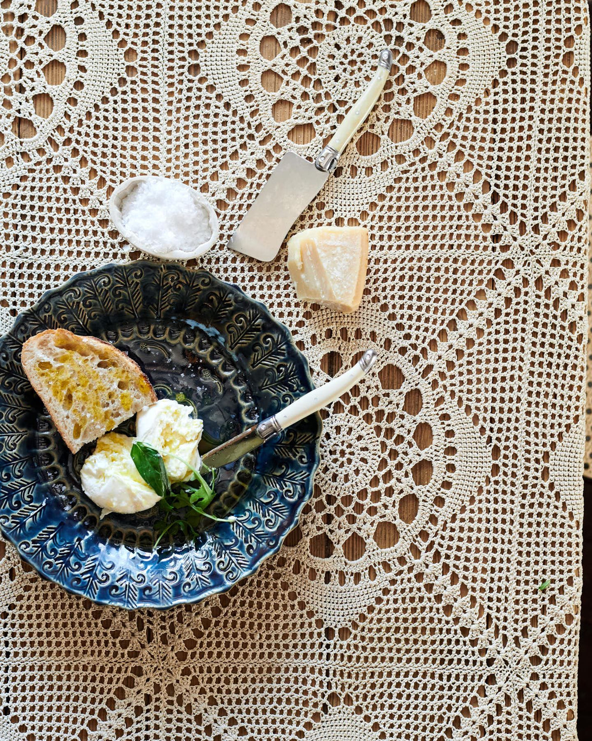 An overhead view of a round handmade bohemian-style blue ceramic platter with vintage silverware, cheese and homemade bread on a beautiful crochet lace tablecloth. TEST