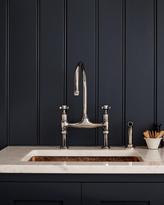 A straight-on view of a traditional-style tarnished silver mixer tap with crosshead handles and an independent rinser tap in a classic utility kitchen with a Carrara marble worktop and deep blue painted cupboards and tongue and groove panelling.