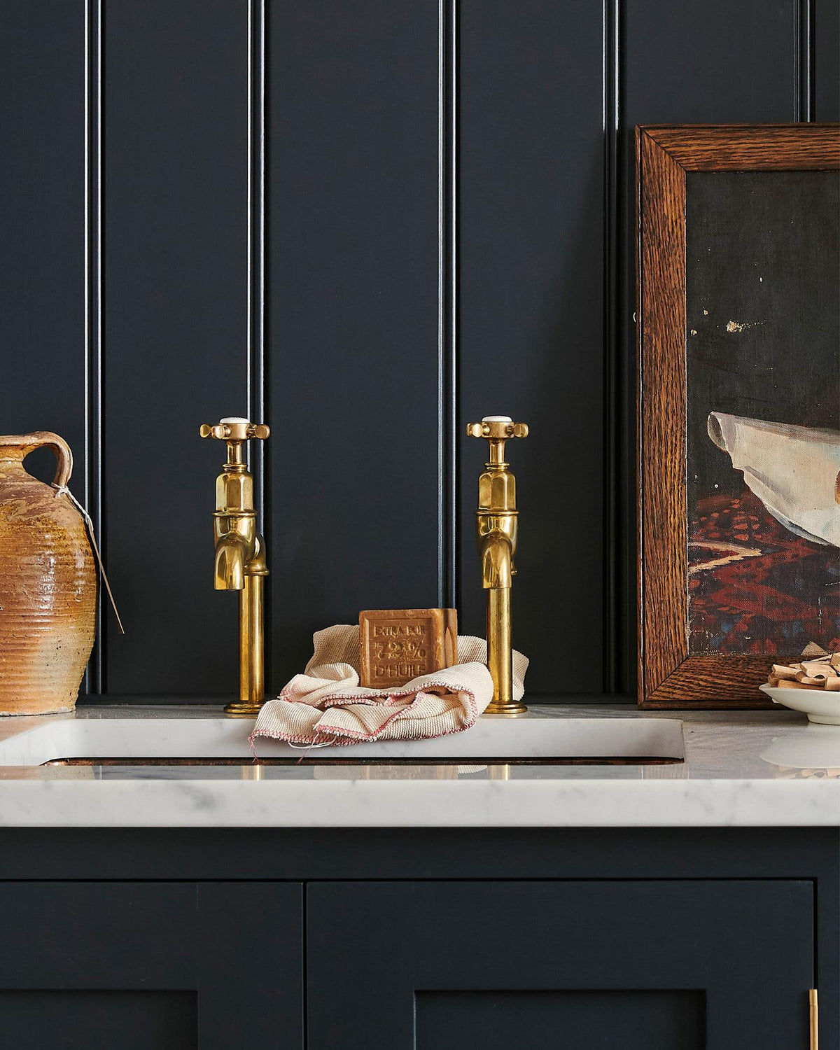 A straight-on view of a pair of traditional-style aged brass bibcock taps with crosshead handles in a classic utility kitchen with a Carrara marble worktop and deep blue painted cupboards and tongue and groove panelling. TEST