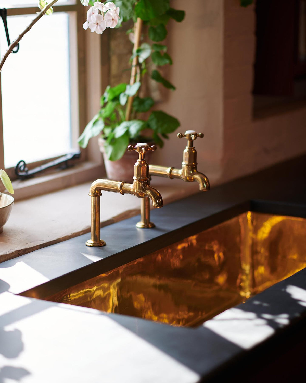 An oblique view of a pair of traditional-style aged brass bibcock taps above a grand polished brass sink and an understated slate worktop. TEST