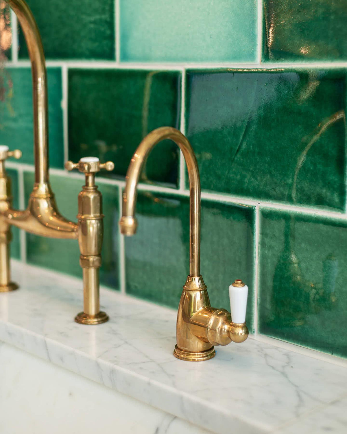 A small traditional-style aged brass tap with a white ceramic lever with glossy emerald green wall tiles behind TEST