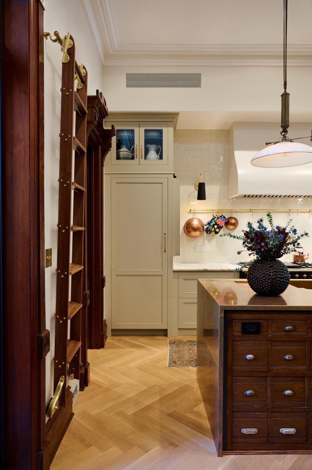 A grand and spacious classic English kitchen features a half vintage and half deVOL kitchen island with a copper sink and worktop, a cooker run with an Arabescato marble worktop, and a full-height cupboard. Beautifully decorated with artisan glass and ceramics, a sophisticated floral arrangement, an aged brass hanging rail, a bespoke ladder, and milk glass pendant lights, it creates a social space with a touch of luxury.