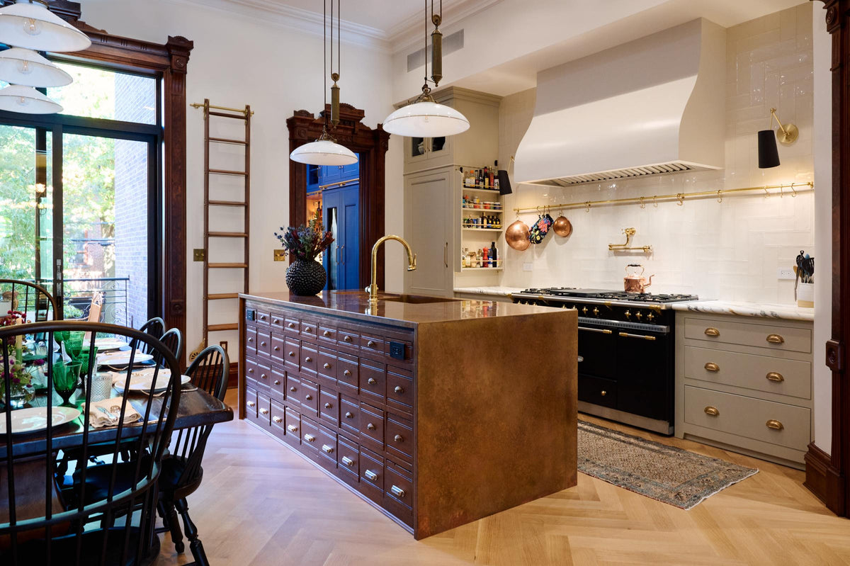 An oblique view of a grand and spacious classic English kitchen features a half vintage and half deVOL kitchen island with a copper sink and worktop. A cooker run with an Arabescato marble worktop and a full-height cupboard. Beautifully decorated with a solid oak dining table set, luscious green house plants, artisan ceramics, a bespoke ladder, an aged brass hanging rail, and milk glass pendant lights, it creates a social space with a touch of luxury.