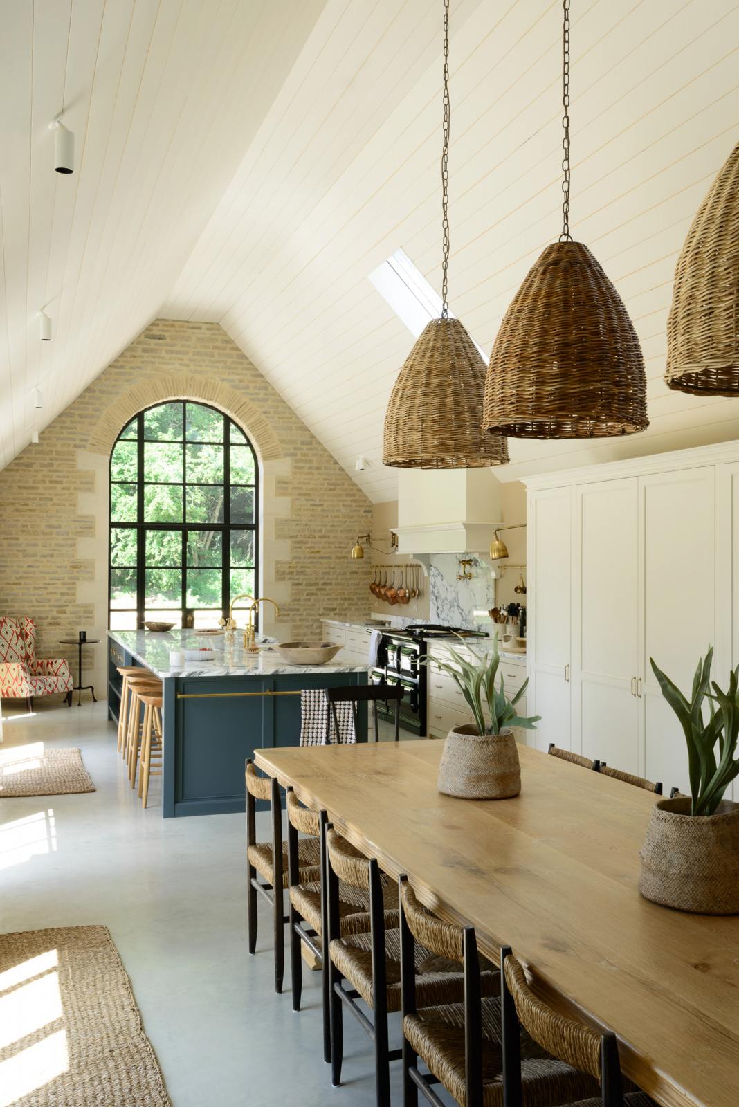 An oblique view of this grand mushroom-coloured classic English kitchen showcases a cooker run with a suspended chimney breast, a large kitchen island topped with Arabescato marble, and a bank of full-height cupboards. Beautifully decorated with a long dining table and woven embellishments such as pendant lights, chairs, rugs, and potted plant covers, this space creates a humble family atmosphere despite its luxurious design.