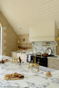 An oblique view of a grand mushroom-coloured classic English kitchen features a cooker run with a suspended chimney breast, a large kitchen island, two sets of sinks, aged brass Ionian taps, and an Arabescato marble worktop. Beautifully decorated with aged brass hanging rails, simple ceramics, brass folded wall lamps, and a large collection of kitchen utensils, this space creates a humble family atmosphere despite its luxurious design.