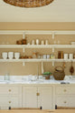 An oblique view of this grand mushroom-coloured classic English kitchen showcases a cooker run with Arabescato marble, a bank of full-height cupboards, and a small sink run with shelves. Beautifully decorated with a long dining table and woven embellishments such as pendant lights, chairs, rugs, a basket, and potted plant covers, this space creates a humble family atmosphere despite its luxurious design.