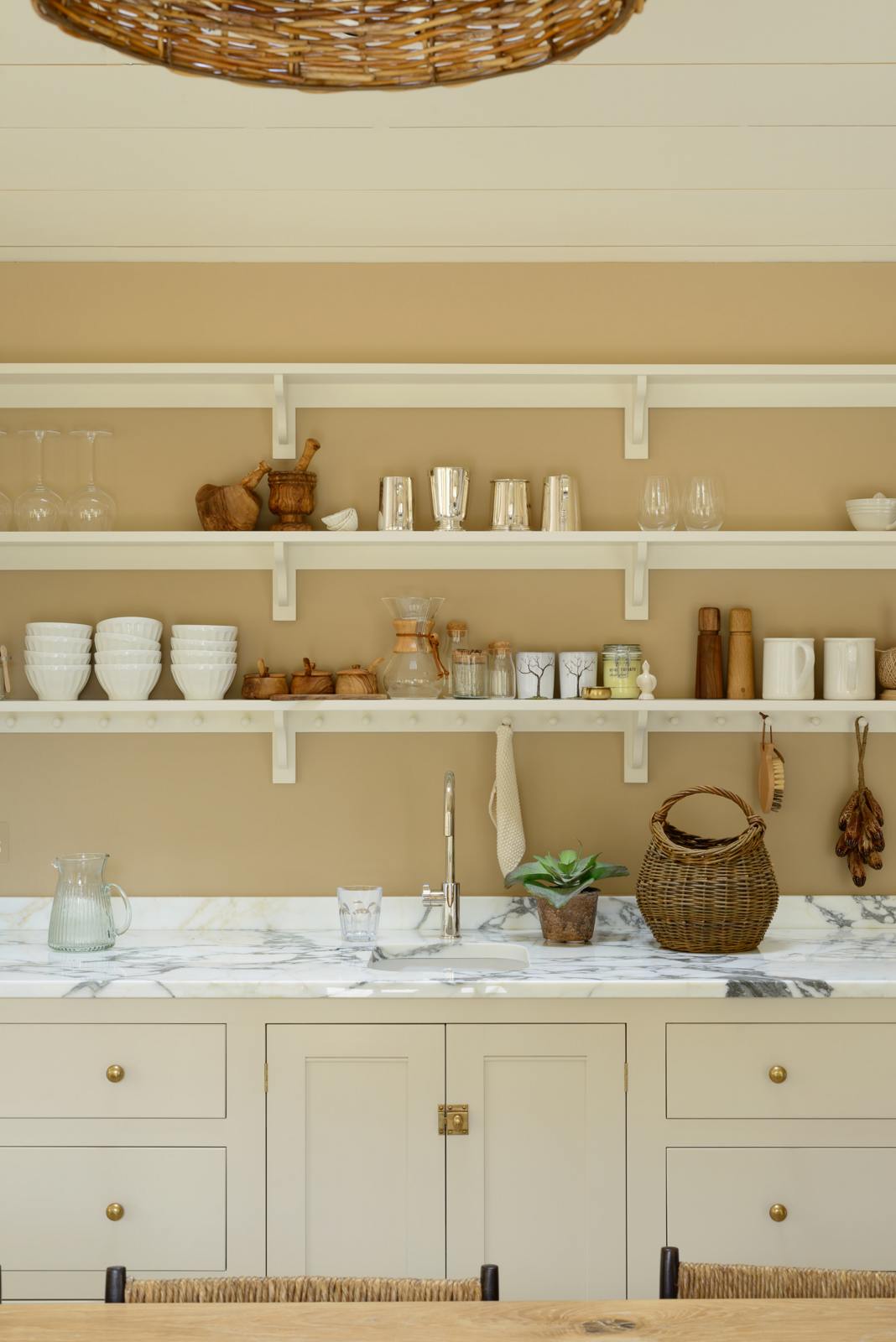An oblique view of this grand mushroom-coloured classic English kitchen showcases a cooker run with Arabescato marble, a bank of full-height cupboards, and a small sink run with shelves. Beautifully decorated with a long dining table and woven embellishments such as pendant lights, chairs, rugs, a basket, and potted plant covers, this space creates a humble family atmosphere despite its luxurious design.