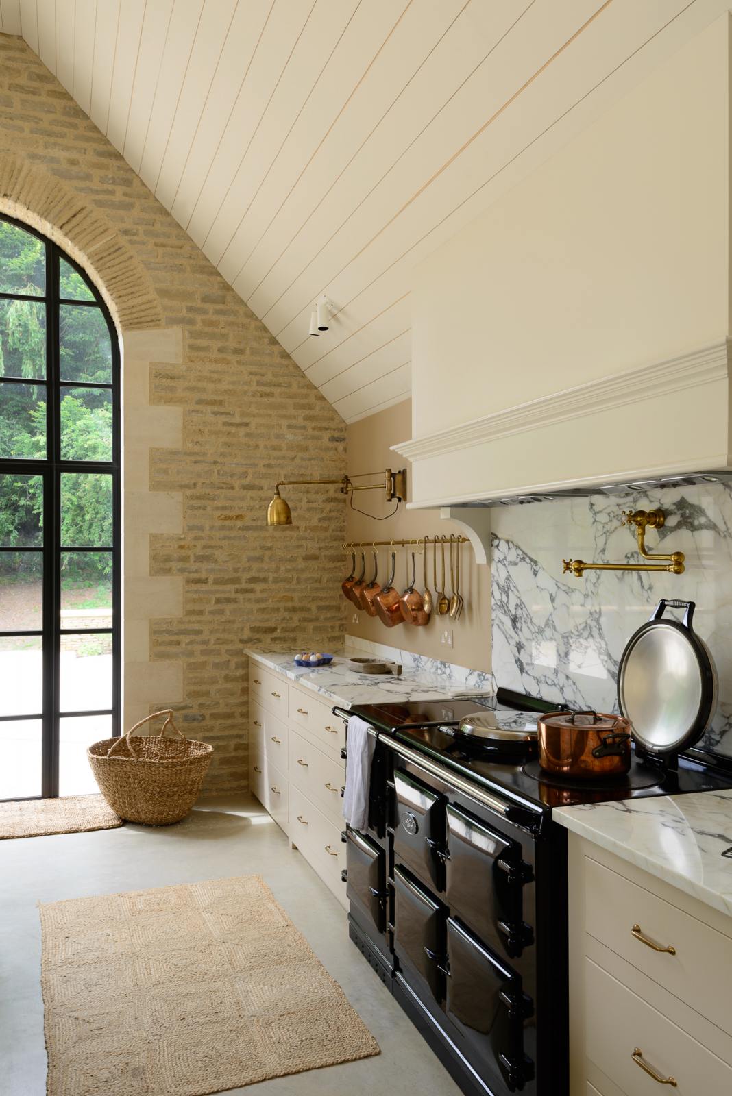 An oblique view of a grand mushroom-coloured classic English kitchen features a cooker run with a suspended chimney breast, a large kitchen island, two sets of sinks, aged brass Ionian taps, and an Arabescato marble worktop. Beautifully decorated with an aged brass hanging rail, copper pans, weaved basket, a brass folded wall lamp, and large gas cooker, this space creates a humble family atmosphere despite its luxurious design.