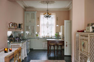 A light pastel-blue classic kitchen and social area features a cooker and sink run with marble worktops, an heirloom dairy table, and Bentwood café stools—colours that oddly fit perfectly into the plaster-pink room, creating a glamorous yet eccentric vibe. The London apartment has high ceilings, worn black painted floorboards, small decorative splashback mirrors, intricate cornices, and is styled with the customer's collection of antiques.