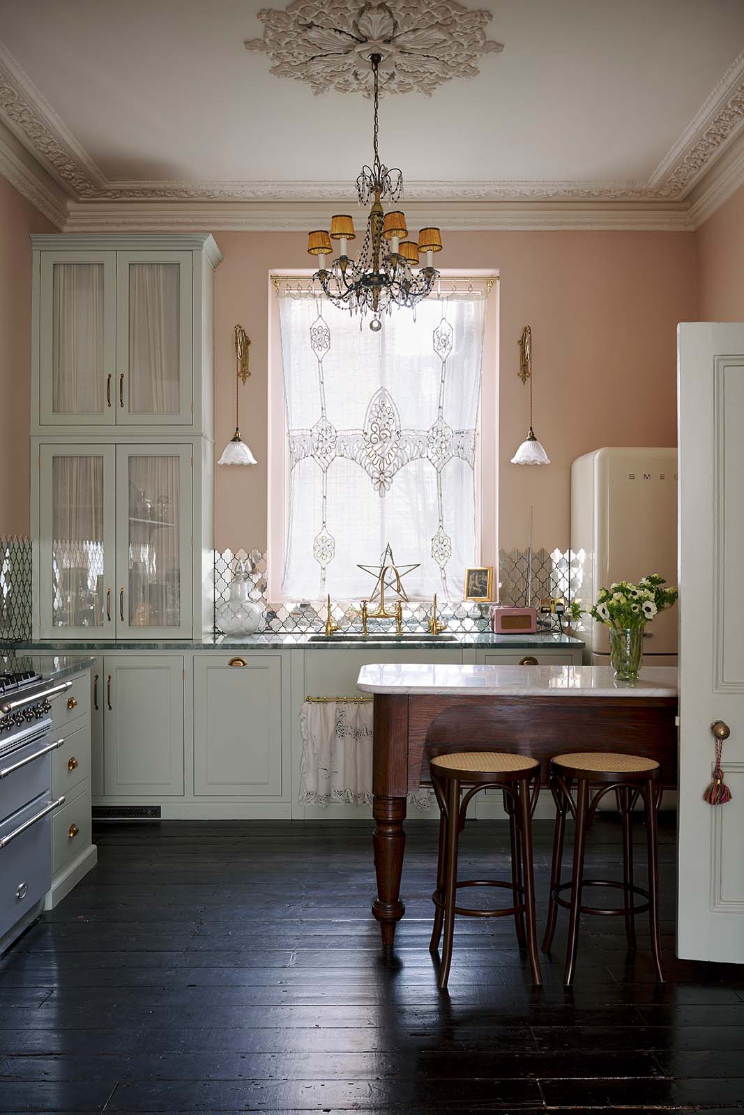 A light pastel-blue classic kitchen and social area features a cooker and sink run with marble worktops, an heirloom dairy table, and Bentwood café stools—colours that oddly fit perfectly into the plaster-pink room, creating a glamorous yet eccentric vibe. The London apartment has high ceilings, worn black painted floorboards, small decorative splashback mirrors, intricate cornices, and is styled with the customer's collection of antiques.