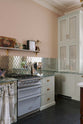 A close and oblique view of the light pastel-blue classic kitchen’s cooker run with a beautiful turquoise marble worktop, and glazed countertop cupboards. The London apartment features high ceilings, worn black-painted floorboards, small decorative splashback mirrors, intricate cornices, and is styled with the customer’s collection of antiques.