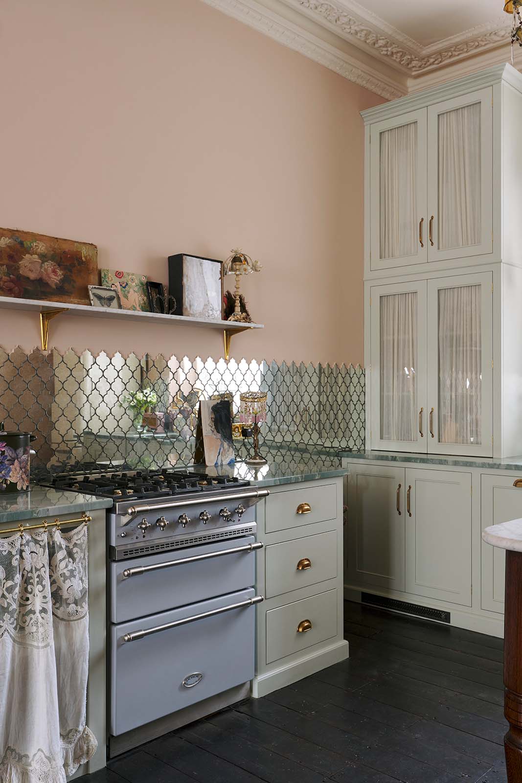 A close and oblique view of the light pastel-blue classic kitchen’s cooker run with a beautiful turquoise marble worktop, and glazed countertop cupboards. The London apartment features high ceilings, worn black-painted floorboards, small decorative splashback mirrors, intricate cornices, and is styled with the customer’s collection of antiques.