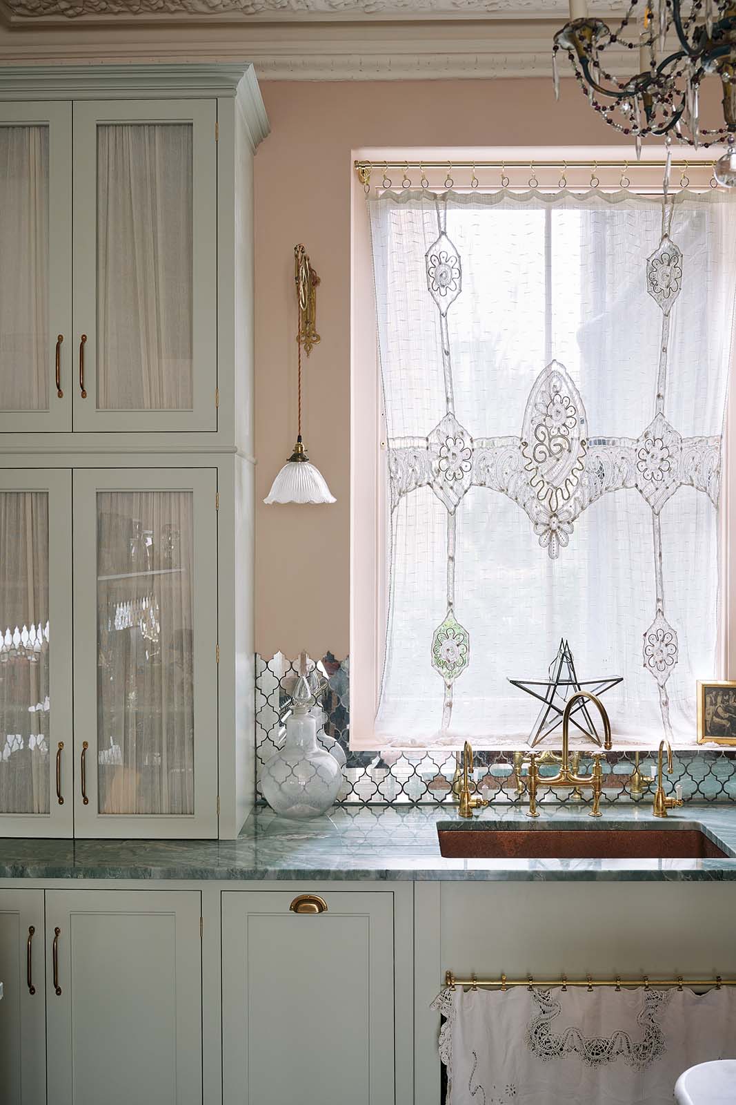 A closer view of the light pastel-blue classic kitchen’s sink run reveals a single copper sink with aged brass Ionian taps and glazed countertop cupboards. The London apartment features high ceilings, worn black-painted floorboards, small decorative splashback mirrors, intricate cornices, and is styled with the customer’s collection of antiques.