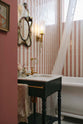 A chic bathroom with pink and white tiled walls, a white bathtub, and a small painter’s black Victorian washtand with aged brass Mayan taps. Above the sink hangs a vintage, eccentric mirror with a highly decorated frame, colourful painted florals, and a feather attached to the side.