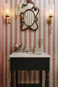 A chic bathroom features pink and white tiled walls, a gold-plated wall light, and a small painter’s black Victorian washstand with aged brass Mayan taps. Above the sink hangs a vintage, eccentric mirror with a richly decorated frame, colourful painted florals, and a feather attached to the side.