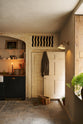 Looking through an open door of a rustic basement kitchen with sandy-coloured walls and ceiling, original Georgian detailing, cream doors and panelling with chipped paintwork and a worn stone floor. On the left is an alcove containing a small run of painted black cupboards with a mottled copper worktop, and a fluted Belfast sink below shelves decorated with vintage bric-a-brac. On the right is an heirloom wall light illuminating.