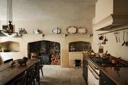 A rustic basement kitchen with sandy-coloured walls and ceiling, original Georgian detailing, cream doors and panelling with chipped paintwork and a worn stone floor. An old banquet-style table sits in the middle of the room with rows of black wooden chairs on either side. On the right is a small cooker run with painted black cupboards with a mottled copper worktop, and a Lacanshe cooker below a devol aged brass hanging rails.