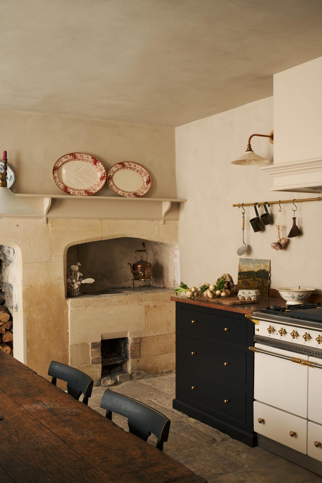 A rustic basement kitchen with sandy-coloured walls and ceiling, original Georgian detailing, cream doors and panelling with chipped paintwork and a worn stone floor. An old banquet-style table sits in the middle of the room with rows of black wooden chairs on either side. On the right is a small cooker run with painted black cupboards with a mottled copper worktop, and a Lacanshe cooker below a devol aged brass hanging rails and heirloom wall light.