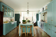 A spacious yet modest Trinity blue classic English kitchen features a Silestone worktop with a sink and cooker run, glazed cupboards, a full-height cabinet with drawers, a kitchen island with a copper sink, aged brass Mayan taps, a copper prep table, and deVOL wooden bum stools, all topped off with a white porcelain splashback. Lovingly decorated with pink peonies in a vase, glass pendant lights, a watermelon, beetroots, live basil, and dried ingredients in glass jars, the room feels humble and warm.