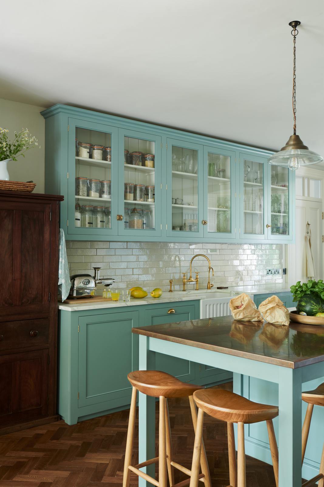 An angled view of a spacious yet modest Trinity Blue classic English kitchen reveals a Silestone worktop sink run with a single fluted Belfast sink and aged brass Ionian tap, glazed cupboards, a copper prep table, and deVOL wooden bum stools—all finished with a white porcelain splashback. Beautifully adorned with wild white flowers in a vase, glass pendant lights, a watermelon, papaya, live basil, dried ingredients in glass jars, the room feels cosy, warm, and inviting.