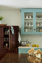 A closer look at a spacious yet modest Trinity Blue classic English kitchen reveals a Silestone worktop sink run, glazed wall cupboards, a copper prep table, a vintage solid oak pantry cupboard, and a white porcelain splashback. Lovingly decorated with a silver juicer, papayas, and dried ingredients in glass jars, the room feels humble, warm, and inviting.